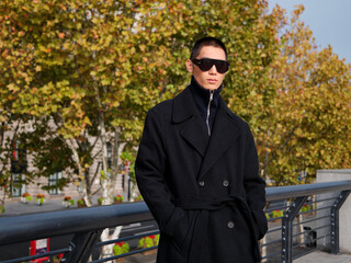 Portrait of handsome Chinese young man wearing black overcoat posing in the street, young guy with black short hair all in black with urban background. Male fashion, cool Asian young man lifestyle.