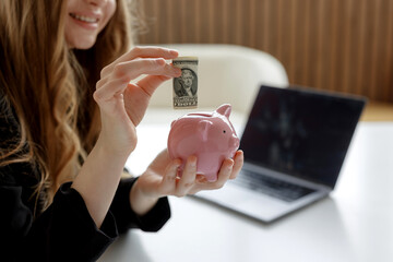 Woman saves dollar bill in pink piggy bank while working at modern office desk
