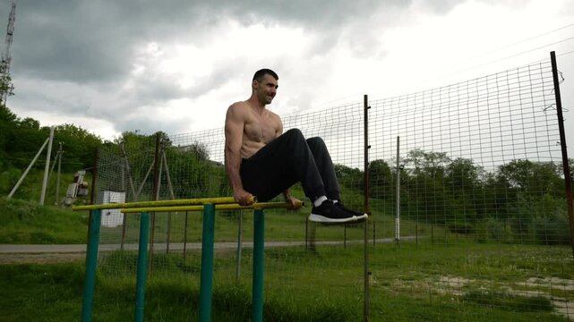 Strong guy training abdominal muscles on parallel bars at sports ground. Young shirtless sportsman exercising at nature. Sporty athlete working out at nature. Concept of active sportive lifestyle