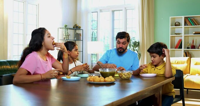 Family eating panipuri snacks together at home, Indian father, mother, son daughter enjoying golgappa at dining table in cozy living room while bonding, chatting, laughing and spending quality time