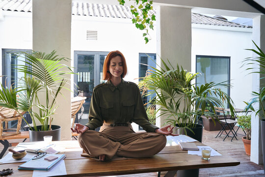 Happy serene young professional business woman, calm mindful lady sitting on work desk meditating, doing yoga breathing exercises for reducing stress in modern green cozy office. Candid photo.