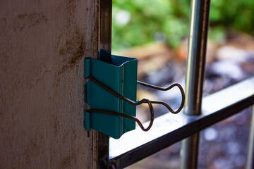 Close-up of a teal binder clip attached to a textured wall near a window with bars and blurred...