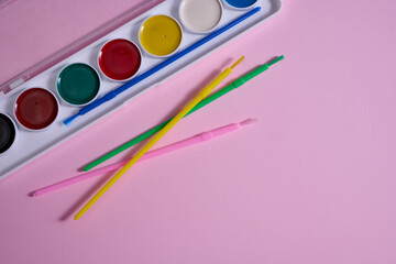 A set of watercolor paints in a plastic case with multi-colored brushes is located on a pink...