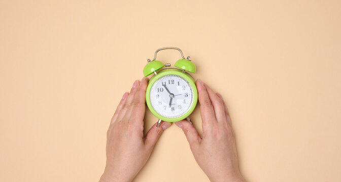 Hands holding green table alarm clock on neutral background.
