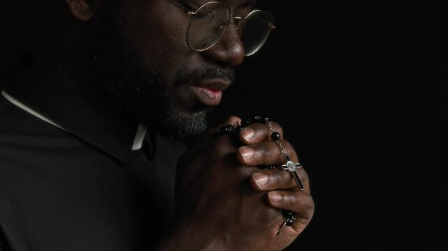 Close-up shot of African American pastor dressed in long-sleeve tonsure shirt murmuring prayer with his eyes closed and hands clasped