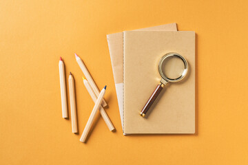 Notebooks with colored pencils and magnifying glass on orange backgroun