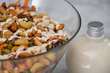 Fresh Broccoli Salad with Cashews in a clear glass bowl with broccoli florets, crunchy cashews, carrots, and red cabbage, drizzled with creamy white sauce bottle closeup view of healthy salad