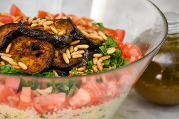 Fresh Mediterranean Couscous Salad with Grilled Eggplant featuring couscous, tomatoes, parsley, grilled eggplant slices, and toasted pine nuts with bottle of olive oil closeup side view