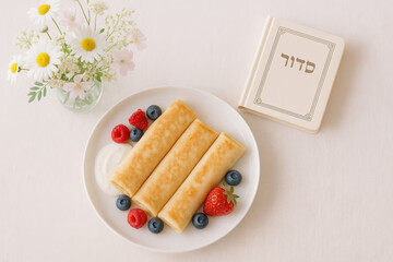 Delicious blintzes with fresh berries and cream on white tablecloth with flowers and prayer book. Shavuot concept.