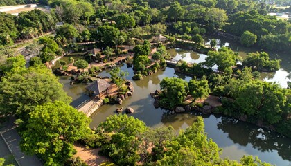 Fototapeta premium Aerial view of a lush, green park with winding paths, ponds, trees, and small structures surrounded by nature.