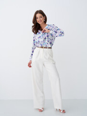 Smiling woman stands confidently in a floral blouse with white high-waisted trousers, hands poised at the collar, wearing heels, styled with long hair and subtle accessories, exuding elegance
