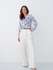 Smiling woman wearing a floral blouse and wide white trousers, hands in pockets. She has brown hair...