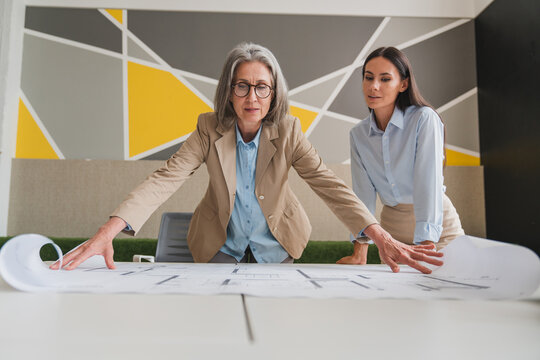 Team of female architects working on construction plans in meeting room. Engineers discussing on project in office. Businesswoman and her colleague standing around table working on blueprint. - Powered by Adobe