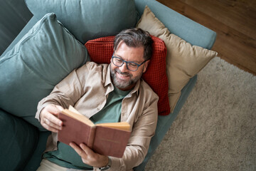 Obraz premium Happy man reading a book while lying on sofa at home