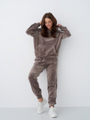 Woman in cozy brown loungewear set with a relaxed pose and white sneakers, adjusting her hair and smiling, standing against a simple white background. Comfortable, casual look