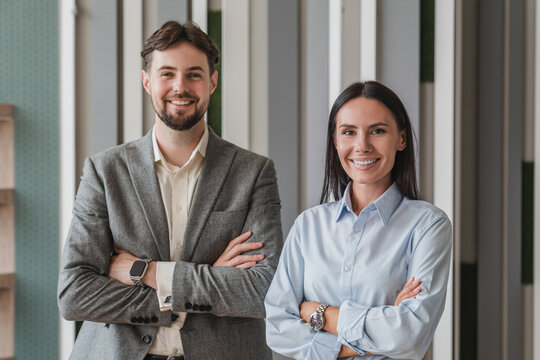 Portrait of smiling young Caucasian business man and business woman standing arms crossed in office looking at camera. Two diverse colleagues, group team of confident professional business people.