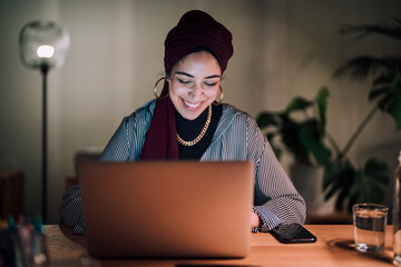 A young Moroccan Arab Muslim woman smiles while working on her laptop at a desk. She is focused and engaged with her work in a home office setting.