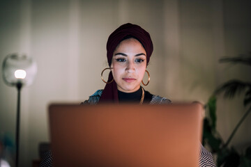 A young Moroccan Arab Muslim woman is focused on her laptop screen while working or studying indoors at night. She concentrates intently on the task.