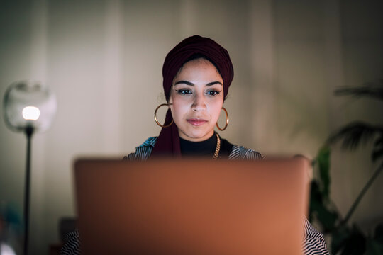 A young Moroccan Arab Muslim woman is focused on her laptop screen while working or studying indoors at night. She concentrates intently on the task.