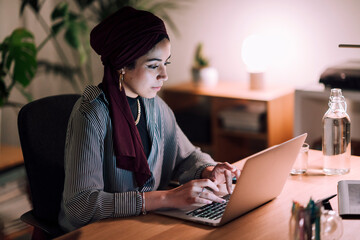 A young Moroccan Arab Muslim woman wearing a hijab works on her laptop at a desk. She is focused on her screen in a dimly lit room.