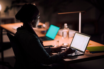 Focused Moroccan Arab Muslim woman working on her laptop in a dark office. She is typing, illuminated by the screen, completing tasks late in the evening.