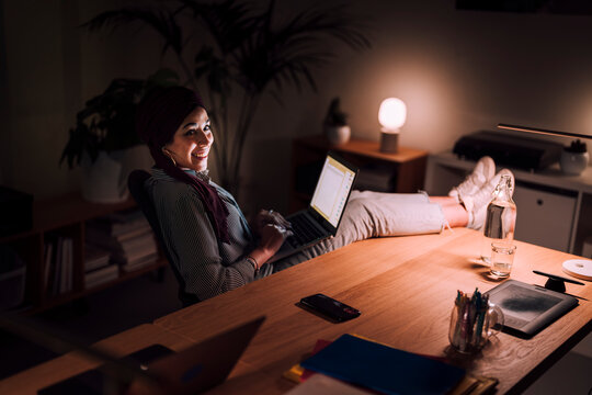 A Moroccan Arab Muslim woman is working late on her laptop in a dark room. She is sitting back comfortably with her feet on the desk, smiling while typing.