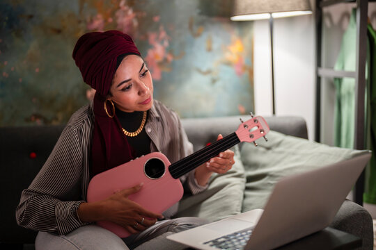 A Moroccan Muslim woman in a turban learns ukulele online. She is Arab and practices music at home with a laptop.
