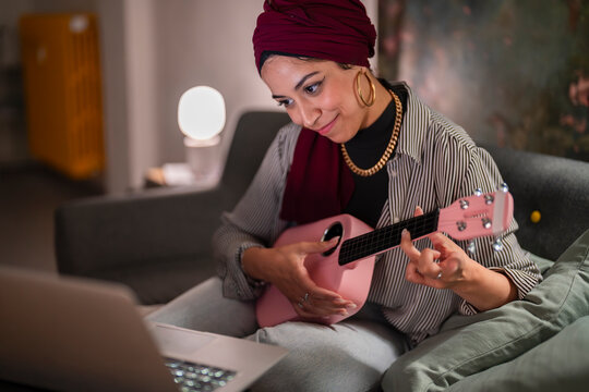 A Moroccan Arab Muslim woman learns ukulele online. She's wearing a turban and sitting on her couch with a laptop, practicing chords on her pink ukulele.