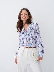 Smiling woman with long hair wearing a floral blouse and white high-waisted trousers, standing with...
