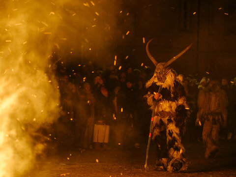 A Krampus figure in a fur suit and horned mask stands amidst smoke and sparks. Krampus and St Nicholas, 5 December in mining village of Cave del Predil, Tarvisio, Italy.