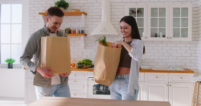 Portrait of happy young couple man and woman coming home from shopping and unpacking paper bags with groceries and vegetables in kitchen together. Healthy food and nutrition concept. 4k video.