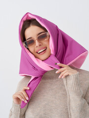 Smiling woman wearing stylish pink hooded scarf and trendy sunglasses, posing with a cheerful expression. She has her hands holding the scarf ends and wears a cozy beige sweater