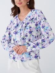 Woman wearing a floral blouse with purple and blue petals, featuring a ruffled front, standing with...