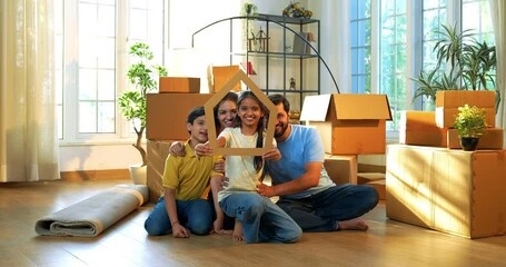 Looking through home shape cutout, Indian family holding paper house model and smiling with joy while shifting to new home, celebrating togetherness, ownership, and new beginnings in modern apartment
