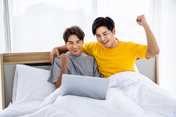 Happy Asian gay couple watching laptop in bed celebrating success together