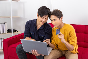 Fototapeta premium Asian LGBT gay couple shopping online with laptop and credit card at home on red sofa