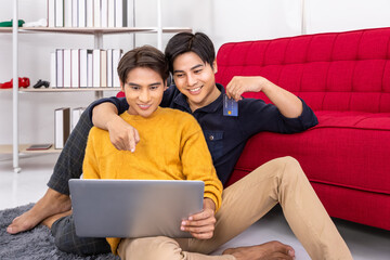 Happy Asian LGBT gay couple shopping online with laptop and credit card while relaxing on the floor