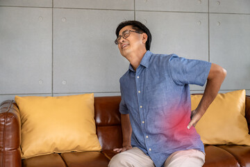 Asian elderly man suffering from lower back pain while sitting on leather sofa