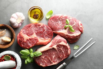 Pieces of raw beef meat, spices and carving fork on grey table, flat lay