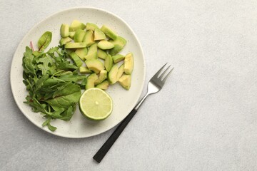 Cut ripe green avocado, greens, lime and fork on light grey table, flat lay. Space for text