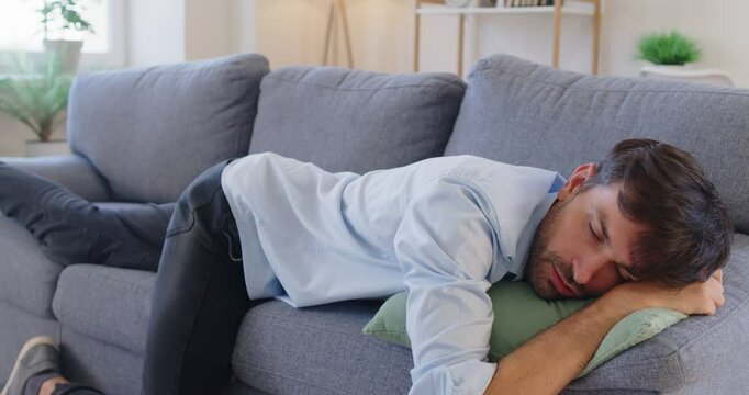 Tired man walking into cozy living room and collapsing onto soft sofa. Exhausted guy lying on couch, showing signs of weariness, resting and seeking comfort and relaxation after long and draining day.