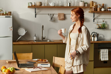 Beautiful red haired woman enjoying coffee in a stylish kitchen while working on a laptop