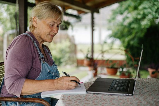 Elderly woman working from home. 
Concentrated good-looking senior woman writing notes in notepad while sitting at table in front of laptop in garden.