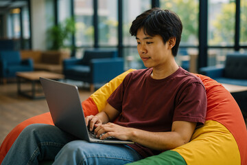 Asian man using laptop while sitting on colorful beanbag chair in modern office space, focused on his work