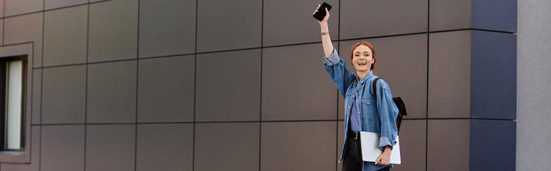 Cheerful red haired woman waves her phone in excitement during a sunny city stroll