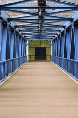 Obraz premium Interior View Of A Modern Covered Pedestrian Bridge With Blue Painted Steel Truss Structure, Concrete Walkway, And Railings. Perspective Leading To End Doors.