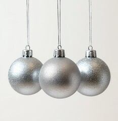 Three silver glittery Christmas baubles hanging on thin silver strings against a plain light background evoking a festive and elegant mood