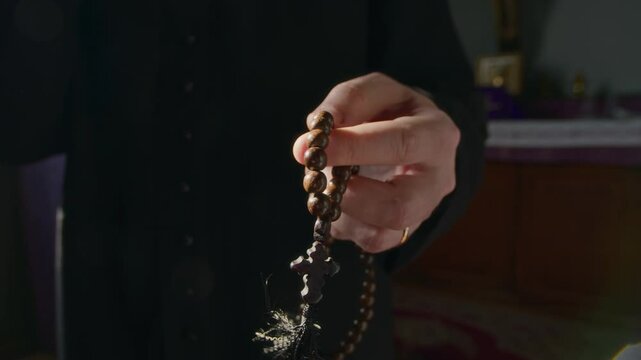 Cropped close up shot of hands of unrecognizable priest in black soutane fingering wooden prayer beads in dim lit church