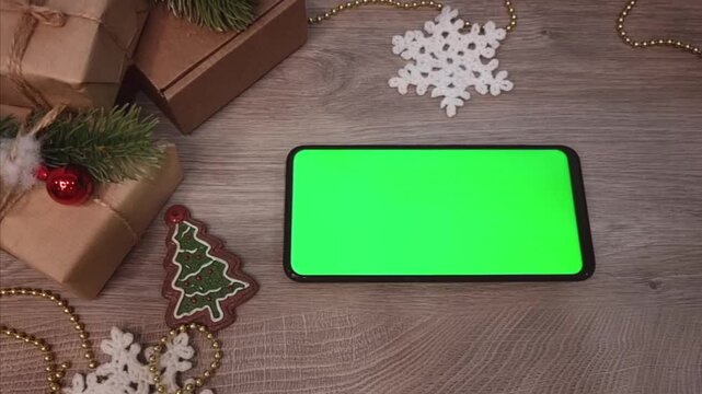 Green screen smartphone lying on wooden table horizontally next to gifts fnd DIY Christmas decorations. Christmas mockup top view.