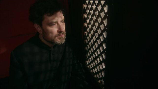 Slowmo shot of middle-aged Caucasian priest in black cassock sitting inside confessional booth and listening carefully to parishioner confession in church